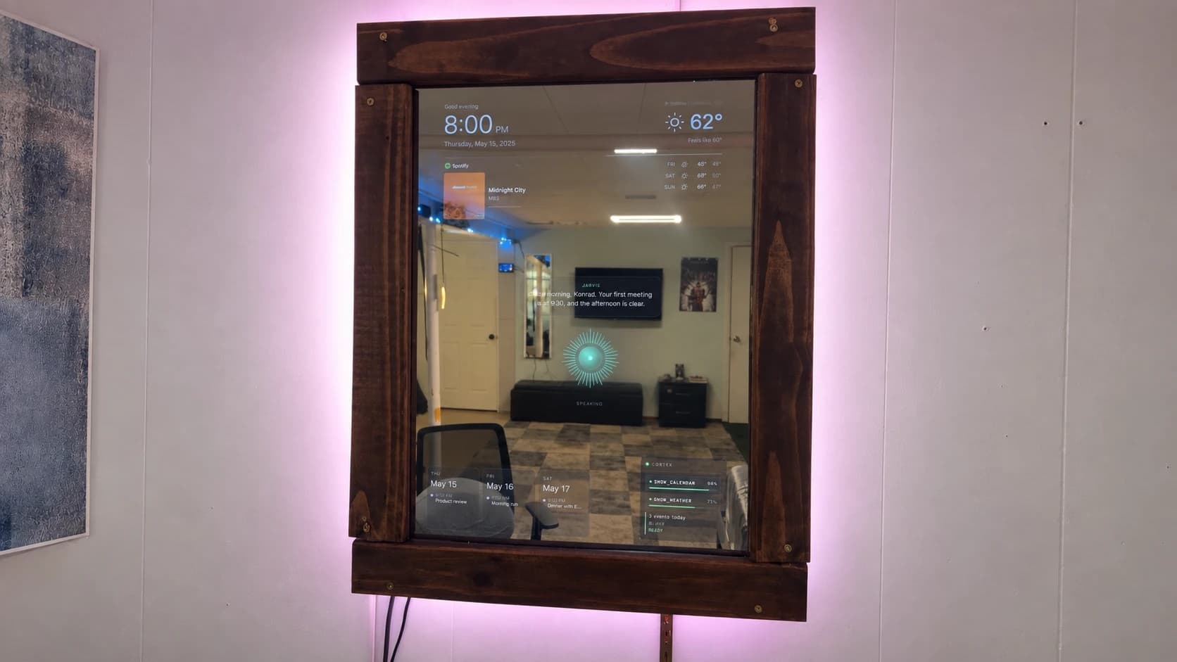 The finished smart mirror on my wall: a dark stained wood frame glowing pink from LEDs behind it, with the clock, weather, Spotify, calendar and a Jarvis reply showing through the glass